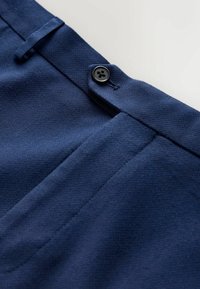 Nahaufnahme des Taillenbunds einer marineblauen Hose mit Knopfverschluss, Gürtelschlaufe und Taschensaum vor hellem Hintergrund.