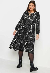 Musta mekko, jossa on valkoinen kukkakuosi, pitkät hihat, avara pääntie ja liehuva hame, yhdistettynä mustiin nilkkureihin.