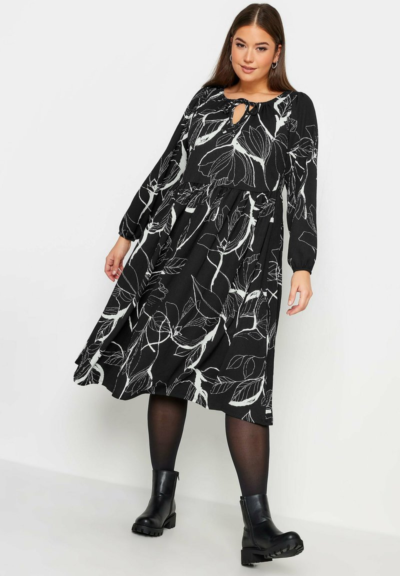 Musta mekko, jossa on valkoinen kukkakuosi, pitkät hihat, avara pääntie ja liehuva hame, yhdistettynä mustiin nilkkureihin.