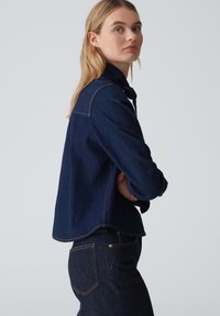 Giacca di denim scuro con un design classico, lunghezza leggermente accorciata, dettagli di cucitura visibili e una vestibilità morbida.