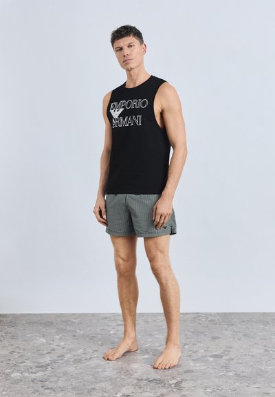Μαύρο μπλουζάκι χωρίς μανίκια με λευκό λογότυπο "Emporio Armani", συνδυασμένο με �γκρι και λευκές ριγέ βερμούδες. Το μοντέλο στέκεται ξυπόλητο σε υφή πατώματος.