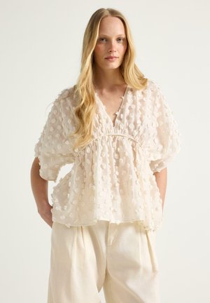 Vaaleahiuksinen nainen, jolla on pitkät hiukset ja päällään kermaisen värinen, teksturoitu paita, jossa on muhkeat hihat, sekä väljät kermaiset housut. Hän seisoo käsivarret selän takana.