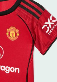 Crvena nogometna majica s crnim detaljima, koja sadrži logo Manchester Uniteda i Adidas pruge na ramenima. Izrađena od lagane tkanine.