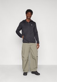 Felpa nera con zip e logo, caratterizzata da una finitura testurizzata e cappuccio. Abbinata a pantaloni larghi di un verde chiaro e scarpe nere.