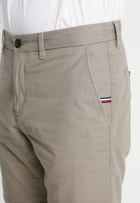 Khakifarbenes kurzes Hosen aus Baumwollmischung mit glatter Textur, ausgestattet mit einem Frontknopfverschluss und Seitentasche, akzentuiert mit einem rot-weiß-blauen Streifen.