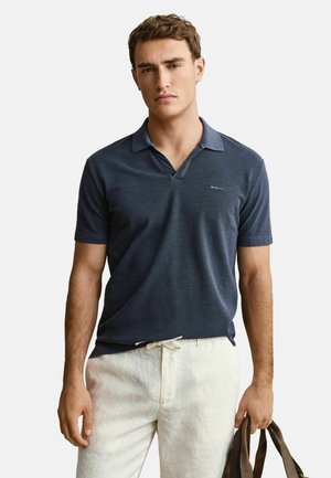 Junger Mann, der ein marineblaues Polohemd und hellbeige Hosen trägt, hält eine braune Tasche in seiner rechten Hand vor einem einfarbigen Hintergrund.