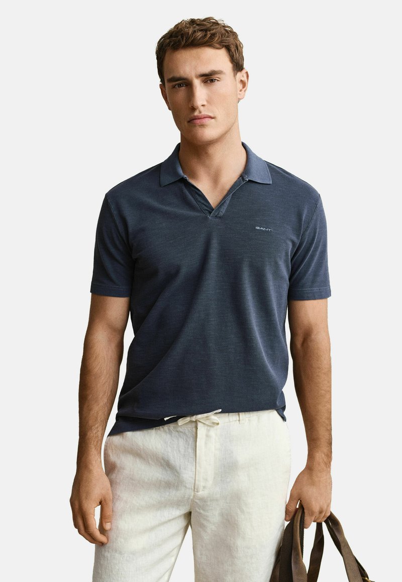 Junger Mann, der ein marineblaues Polohemd und hellbeige Hosen trägt, hält eine braune Tasche in seiner rechten Hand vor einem einfarbigen Hintergrund.