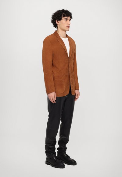 Brown suede blazer with lapels, single button closure, two front pockets. Paired with black leather pants and shoes, standing against a plain background.
