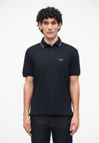 Čierne polo tričko s priliehavým strihom, obsahujúce troj gombíkovú légou, biele lemovanie goliera a malé logo na ľavej hrudi.