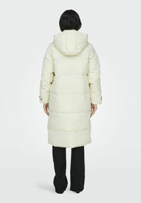 Abrigo puffer largo en blanco roto, con capucha, acolchado en horizontal y bolsillos laterales. Llevado sobre pantalones negros.