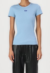 Kobieta ubrana w dopasowaną, jasnoniebieską, krótką koszulkę z prążkowanego materiału z małym logo "HUGO" na piersi oraz luźne, czarne skórzane spodnie.