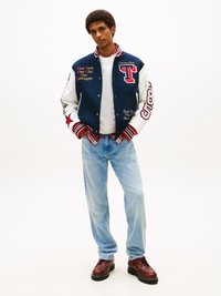 Chaqueta varsity azul y blanca con texto y gráficos bordados, combinada con una camiseta blanca y jeans azul claro. Zapatos marrones con cordones.
