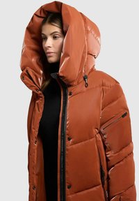 Cappotto imbottito arancione con un ampio cappuccio, zip frontale e tasche laterali. Il materiale appare lucido con una texture trapuntata. Accenti neri sull'hardware.