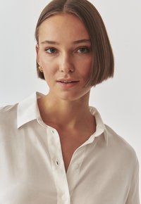 Bílá košile na knoflíky, hladká textura, klasický límeček, viditelný jeden knoflík. Model má krátké vlasy a minimální doplňky. Jednoduché pozadí.