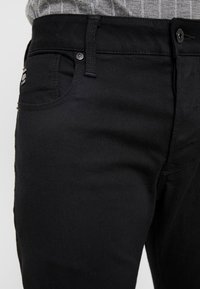 Svarta jeans i denim med raka ben, utrustade med en framficka, metallknappsförslutning och subtil branding på midjebandet.