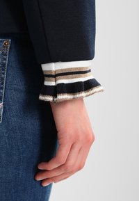 Granatowy sweter z prążkowanym mankietem w paski w kolorze białym, złotym i czarnym. Mankiet ma falbaniasty detal. Noszony z niebieskimi dżinsami.