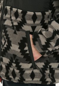 Fleecejacke mit einem schwarz-grauen Tribal-Muster, ausgestattet mit einer Reißverschlusstasche und einem kontrastierenden schwarzen Einsatz im oberen Bereich. Weiche Textur.