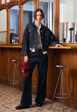 Giovane con capelli corti e scuri, indossa una giacca di jeans nera e pantaloni, tiene in mano un berretto rosso e si appoggia a un bancone di legno con sgabelli in metallo.