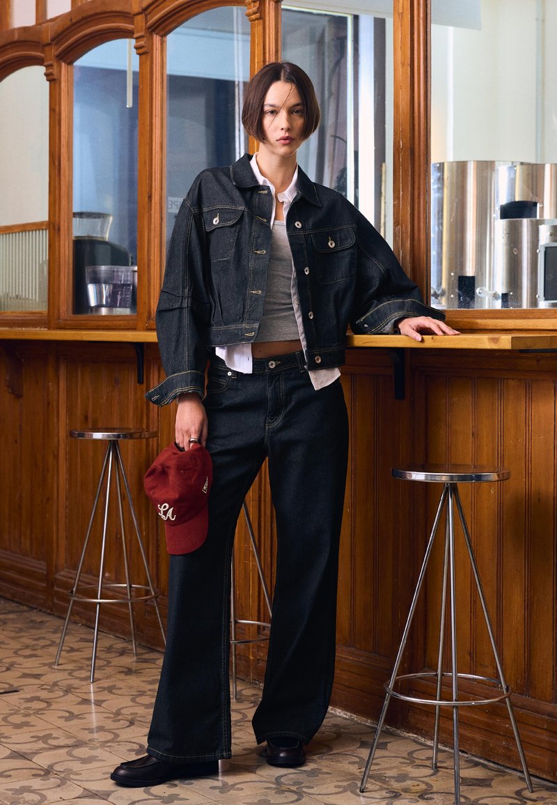 Fiatal személy rövid, sötét hajjal, fekete farmerkabátban és nadrágban, piros sapkát tartva, fának támaszkodva, fém székekkel körülvéve.