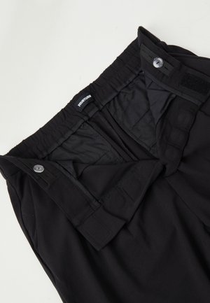 Fascia dei pantaloni neri con elastico regolabile e chiusure a bottone, parzialmente aperti per mostrare l'etichetta interna e i dettagli della chiusura.