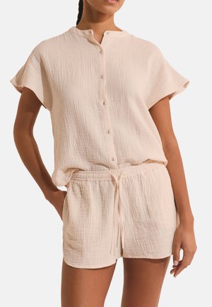 Kobieta ubrana w jasno-beżową, teksturowaną koszulę na krótki rękaw z guzikami i dopasowane szorty ze sznurkiem, stojąca z jedną ręką w kieszeni.