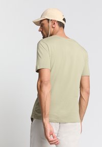 Licht olijfgroen T-shirt met korte mouwen en ronde hals, gladde textuur en een relaxte pasvorm. Gekozen in combinatie met een lichtbeige pet.