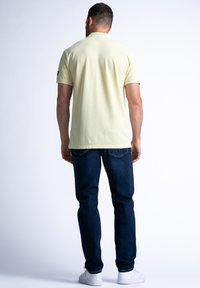 Koszulka polo w jasnożółtym kolorze, z krótkim rękawem, wykonana z miękkiej bawełny. Połączona z ciemnoniebieskimi dżinsami i białymi trampkami, widok z tyłu.