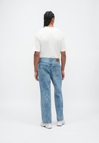 Mężczyzna z warkoczami, ubrany w biały t-shirt, niebieskie dżinsy w stylu acid-wash i białe trampki, stojący tyłem na jednolitym białym tle.