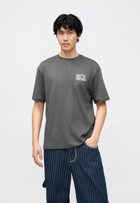 Siva majica kratkih rukava s bijelim natpisom "SRETNI BLagdani" na lijevoj strani prsa, uparena s tamnoplavim hlačama s tankim bijelim prugama.