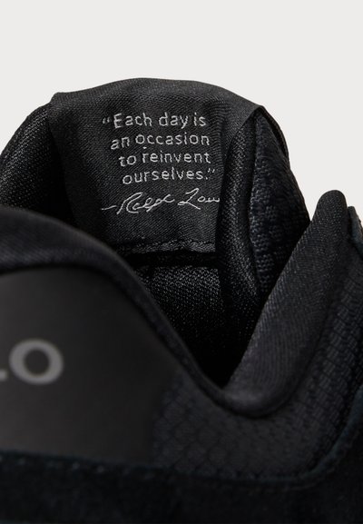 Crni unutrašnjost tenisice s teksturiranom tkaninom, s bijelom naljepnicom s citatom koja je prišivena. Gornji rub ima glatku završnu obradu i okrugli oblik.