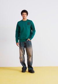 Grønn bomullssweatshirt med en svart lomme, kombinert med slitte blå jeans. Modellen står på et gult gulv foran en hvit bakgrunn.