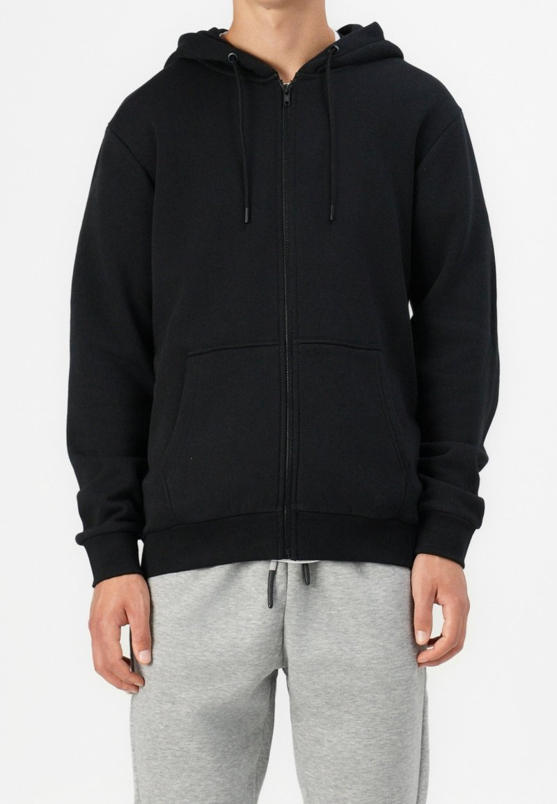 Sudadera con cremallera negra y un bolsillo frontal, con cordones y puños acanalados. Llevada sobre pantalones de chándal grises, con un diseño sencillo y de color sólido.