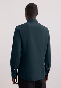 Camicia a maniche lunghe color verde scuro con colletto standard, polsini con bottone e una consistenza liscia. Presenta un orlo dritto e una morbida caduta.