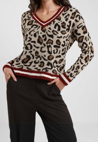Maglione leopardato in beige, con scollatura a V, polsini e orlo a coste a righe rosse e bianche. Morbido tessuto a maglia. Abbinato a pantaloni scuri.