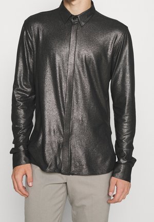 Homme portant une chemise noire métallisée à manches longues avec col et boutons, associée à un pantalon beige clair, debout devant un fond uni.