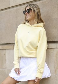 Geel katoenen hoodie met een logo aan de voorkant, gecombineerd met een witte rok. De hoodie heeft een losse pasvorm en lange mouwen. Zachte textuur.