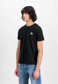 T-shirt de algodão preto com mangas curtas, decote redondo e um pequeno logótipo branco no peito. Usada com jeans de denim azul claro.