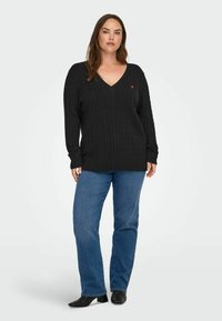 Black cable knit sweater with a V-neck, featuring long sleeves and a small red heart logo. Worn with blue straight-leg jeans and black ankle boots.