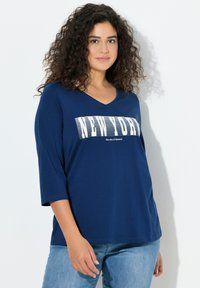 Marineblaues V-Ausschnitt-Shirt mit dreiviertellangen Ärmeln, mit einem weißen Grafikdruck, der "NEW YORK" und "die Stadt der Träume" zeigt. Weiche Stoffstruktur.