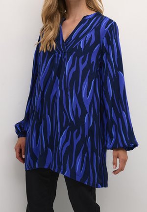 Vrouw draagt een loszittende tuniek met lange mouwen, met een blauw-zwart abstract tijgerstreepjespatroon en een V-hals.