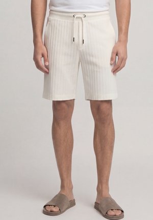 Mann trägt cremefarbene, gerippte Shorts mit Kordelzug, kombiniert mit taupefarbenen Slide-Sandalen, steht vor neutralem Hintergrund.