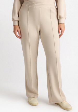 Pantalons larges beige en tissu lisse et extensible avec des coutures latérales, une taille élastique et une coupe décontractée. Associés à des baskets de couleur claire.