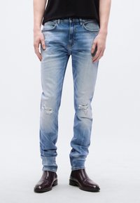 Personne portant un jean délavé bleu clair et des bottines en cuir marron foncé, debout devant un fond clair uni.