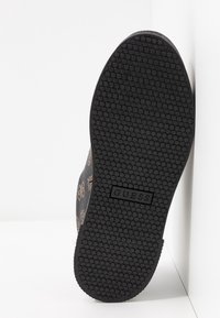 Semelle d'une chaussure noire avec motif de bande de roulement circulaire et logo "GUESS" au centre, inclinée sur fond blanc.