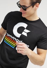 Camiseta negra con un gran gráfico blanco de la letra "C" y el texto estilizado "commodore", con un patrón de franjas horizontales multicolores.