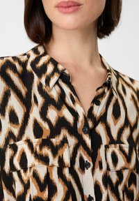Leopardenmuster-Bluse mit einem schwarz, beige und braun gemusterten Design, mit Kragen, Knopfleiste und Brusttaschen. Weicher Stoff.