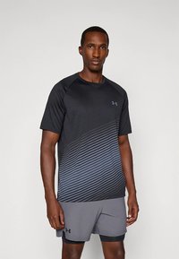 Črna atletska majica s sivo prelivajočim vzorcem črt, kratkimi raglan rokavi in logotipom Under Armour na prsih, kombinirana s sivimi kratkimi hlačami.