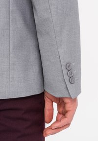 Harmaa bleiserin hiha, jossa on teksturoitu kangas, kolme nappikoristetta ja hillityt saumat. Kädet lepäävät tumman marooninväristen housujen päällä.