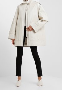 Krämfärgad oversized kappa med hög krage, dragkedjestängning och stora fickor. Har en slät textur och framträdande knappdetaljer.