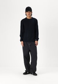 Suéter negro de punto trenzado con diseño texturizado, combinado con jeans negros de corte holgado y zapatillas negras, llevado con una gorra de béisbol negra.